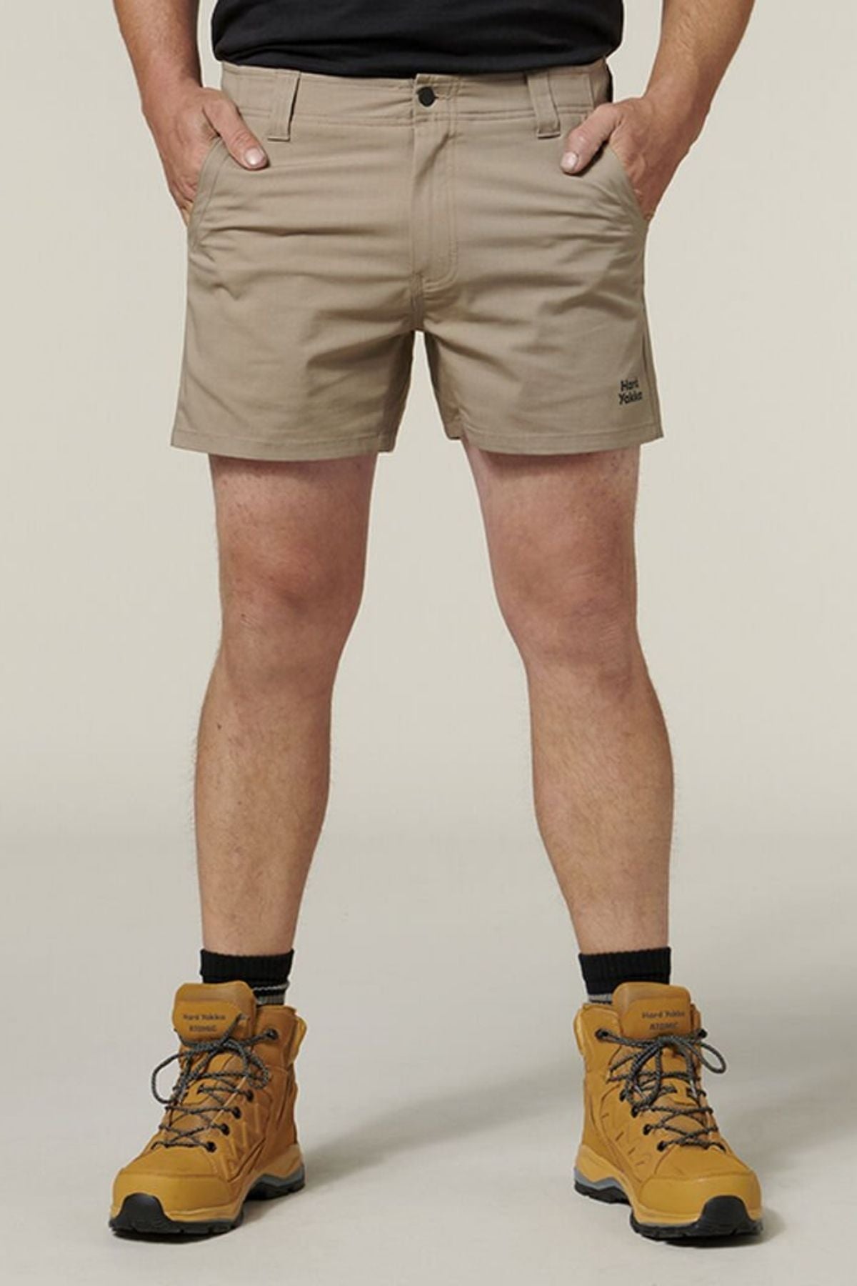A person wears beige Hard Yakka Raptor Short Shorts with Sustainable REPREVE® ECO Ripstop Technology and tan hiking boots. The shorts feature an Active Fit waistband and a logo on the left leg. The background is plain off-white.