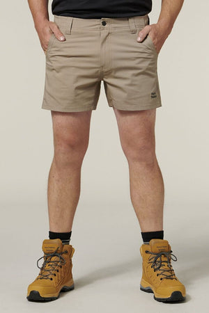 A person wears beige Hard Yakka Raptor Short Shorts with Sustainable REPREVE® ECO Ripstop Technology and tan hiking boots. The shorts feature an Active Fit waistband and a logo on the left leg. The background is plain off-white.
