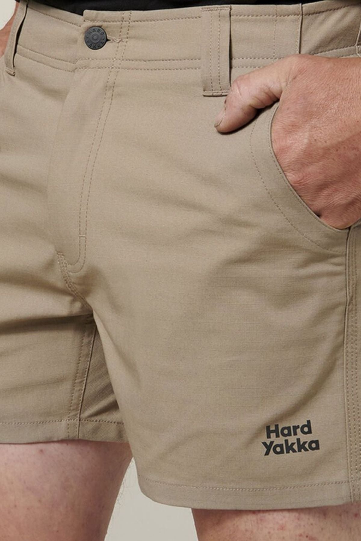 Close-up of a person wearing beige Hard Yakka Raptor Short Shorts made from sustainable REPREVE® fabric, with one hand in the pocket. The Hard Yakka logo appears on the lower left leg.