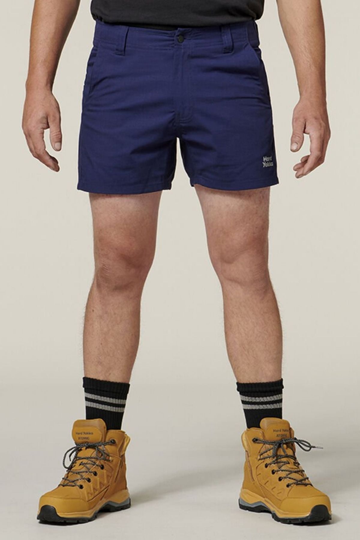 A person is wearing the comfortable Hard Yakka Raptor Short Shorts in dark blue, which have the words "Hard Yakka" on the left leg, along with a black shirt tucked in. The shorts feature an Active Fit waistband for added ease. The ensemble is completed with brown hiking boots and black socks with white stripes, set against a plain, light-colored background.