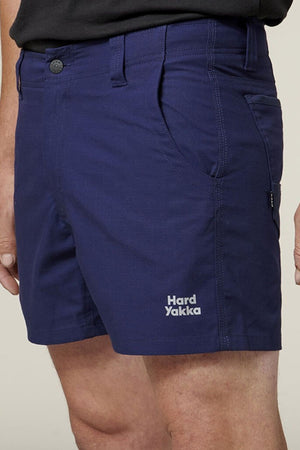 Close-up of a person wearing dark blue Hard Yakka Raptor Short Shorts. These comfortable shorts feature front utility pockets and a button closure, with the Hard Yakka logo printed in white on the lower left side. The person is wearing a black shirt tucked into the shorts.