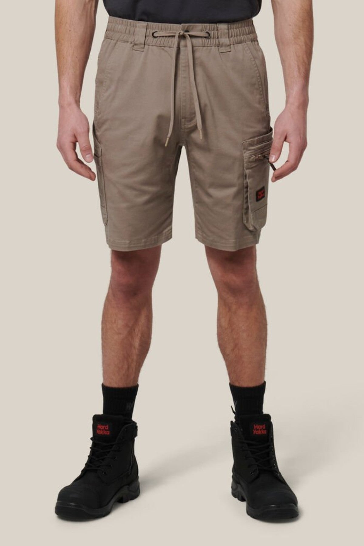 A person wearing Hard Yakka Tough Maxx Mid Shorts—made from durable canvas with UPF 50+—along with black work boots and socks, stands before a beige background. The shorts have multiple pockets and a drawstring waistband.