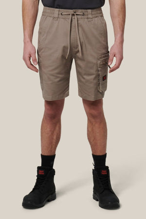 A person wearing Hard Yakka Tough Maxx Mid Shorts—made from durable canvas with UPF 50+—along with black work boots and socks, stands before a beige background. The shorts have multiple pockets and a drawstring waistband.