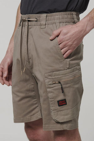 A person wears Hard Yakka Tough Maxx Mid Short cargo shorts in light brown, featuring an elastic waistband, drawstring, multiple pockets including a zipped side pocket with a Hard Yakka patch, and durable canvas fabric. One hand is in the front pocket.