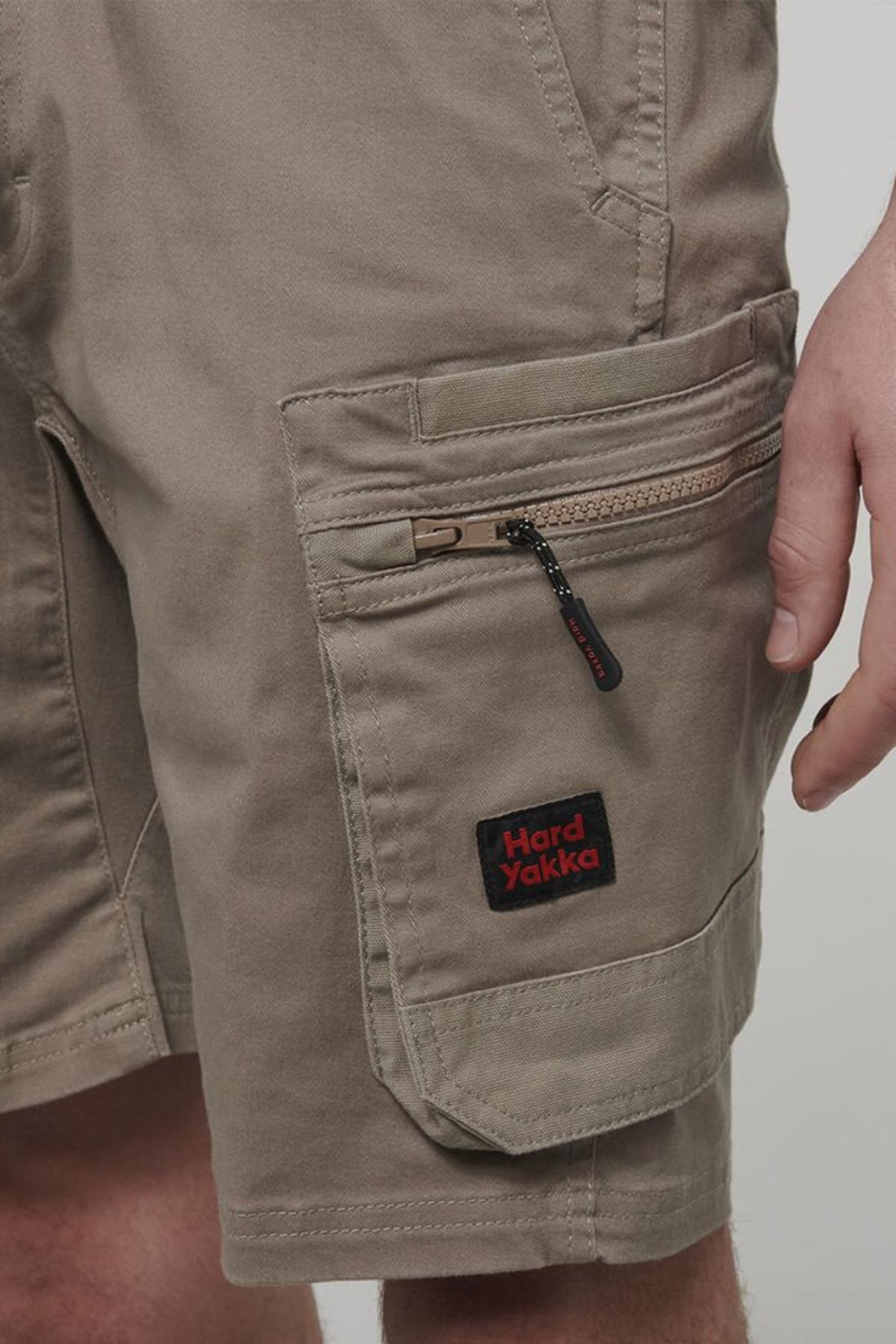 A close-up shows a person wearing Hard Yakka Tough Maxx Mid Short khaki shorts with durable canvas, zippered side pocket, and a black and red "Hard Yakka" logo patch. Only part of the hand and upper leg are visible.