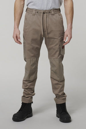 A person stands facing forward, wearing Hard Yakka Tough Maxx Pants with multiple pockets, ankle cuffs, and a drawstring waist. Black lace-up boots and a partially visible gray T-shirt complete the look; upper body is mostly out of frame.