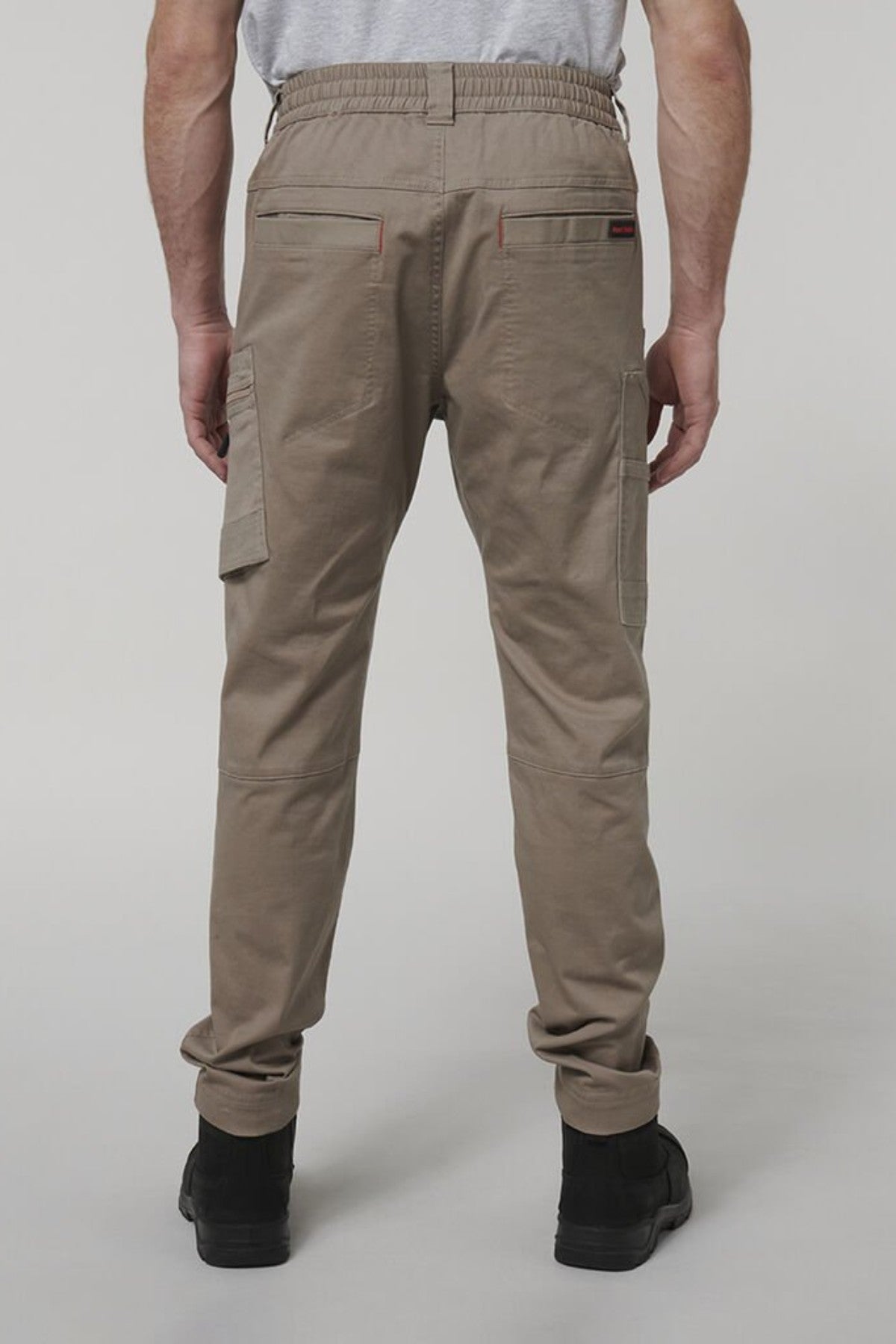 A person stands facing away in Hard Yakka Tough Maxx Pants—light brown, multi-pocketed cargo pants with rear flap pockets—paired with a gray T-shirt and black boots. The background is plain and light-colored.