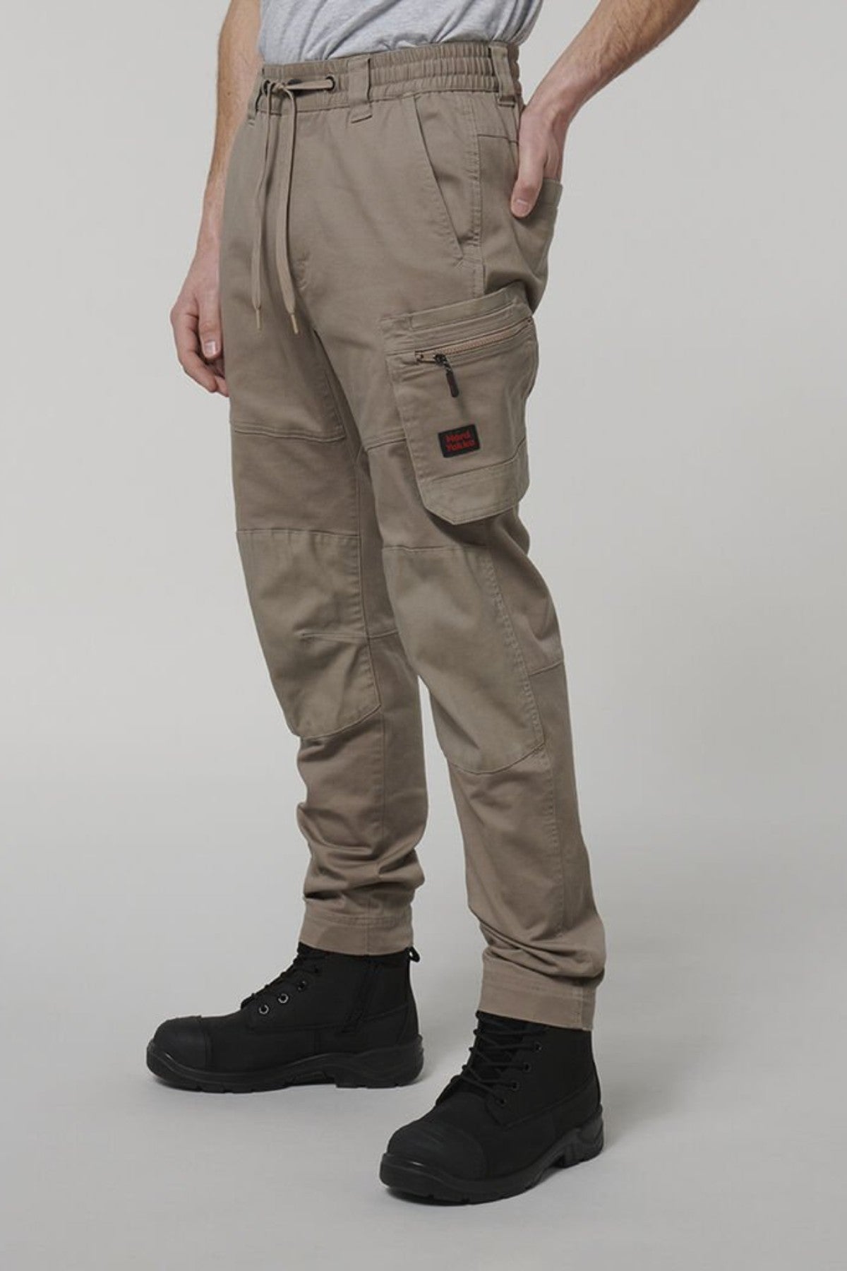 A person wears Hard Yakka Tough Maxx Pants in light brown with multiple pockets and black boots, standing against a plain light gray background. The upper body is mostly out of frame.