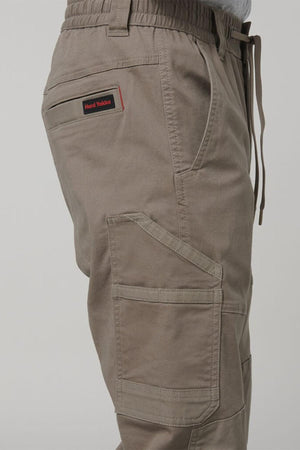 Side view of a person wearing Hard Yakka Tough Maxx Pant cargo pants featuring multiple pockets and a drawstring waistband. A red and black "Hard Yakka" label is visible above the left thigh pocket.
