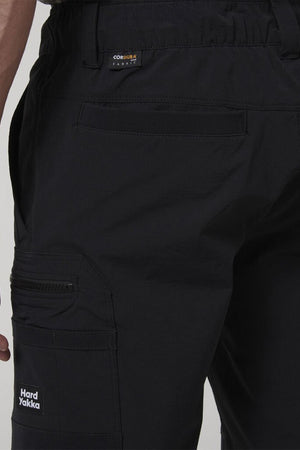Close-up of a person wearing Hard Yakka X Range Mid Short in black, made from durable workwear materials with multiple pockets and zippers. Visible labels show "Hard Yakka" on the thigh and "CORDURA FABRIC" near the waistband.