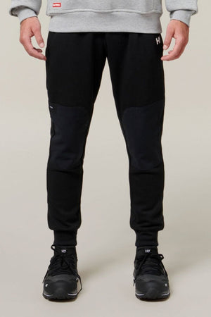 A person stands facing forward, wearing a grey sweatshirt, Hard Yakka Xtreme Jogger black track pants with reinforced knee patches, and black sneakers. Only the lower half of the body is visible against a plain background.