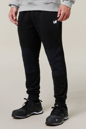Against a neutral background, only the lower half of an individual is visible as they wear black Hard Yakka Xtreme Jogger pants featuring reinforced knee patches and a small white "HY" logo on the left thigh. They also sport black sneakers and a gray sweatshirt.