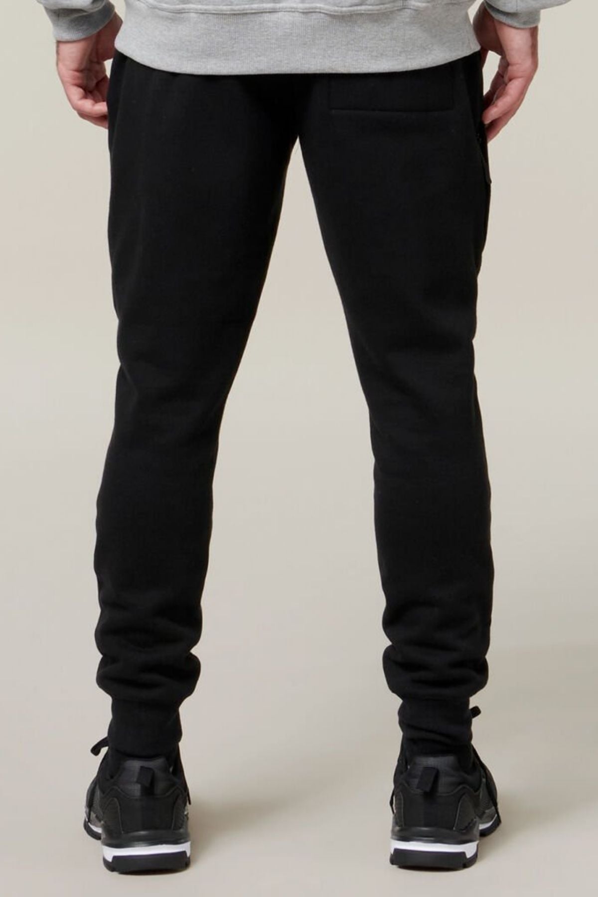 The image shows a person standing and facing away from the camera, wearing Hard Yakka Xtreme Jogger pants with reinforced knee patches and black sneakers. The gray waistband of a Hard Yakka hoodie or sweatshirt is visible, and the setting appears to be indoors with a plain, light-colored background.