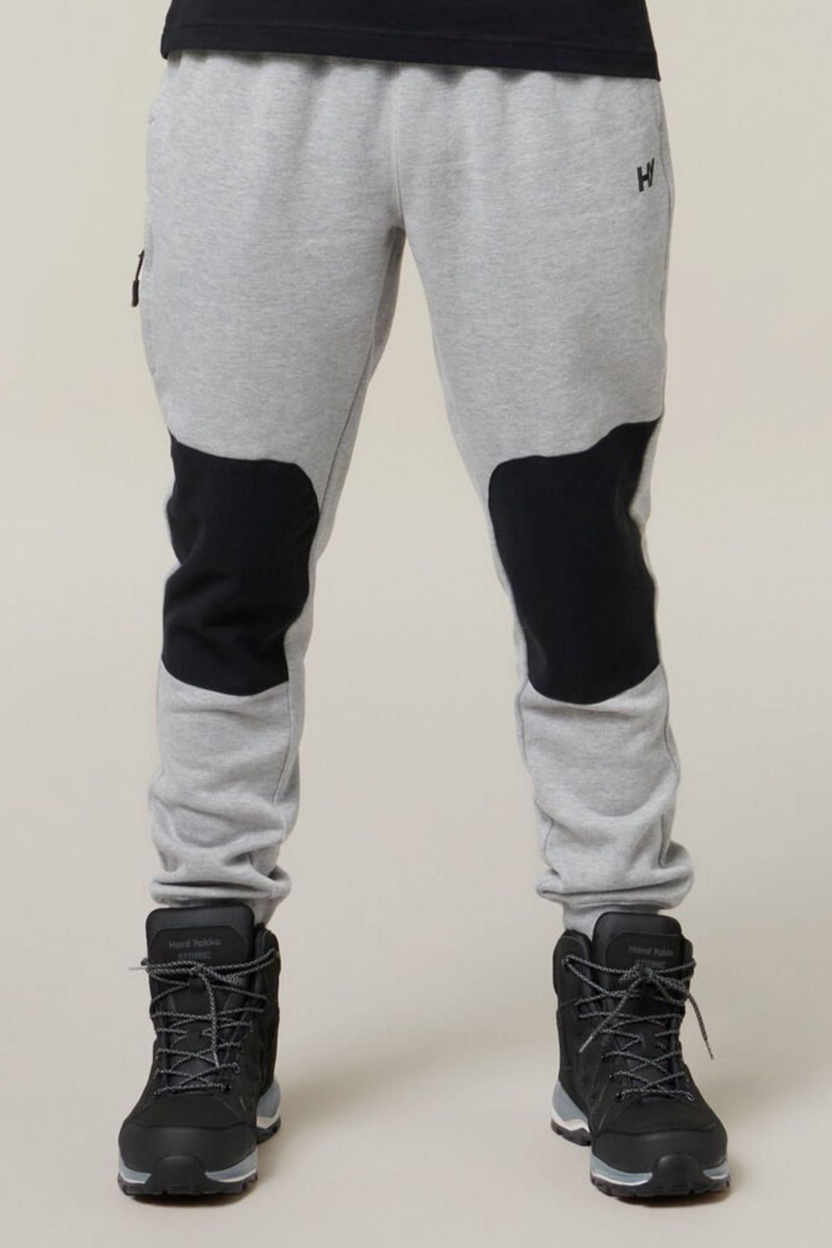 A person wearing Hard Yakka Xtreme Jogger pants with reinforced knee patches and black high-top sneakers. The background is plain beige, and only the lower half of the person's body is visible.