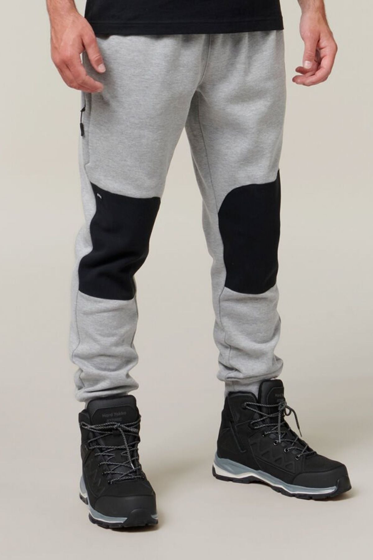A person wearing a pair of Hard Yakka Xtreme Joggers with reinforced knee patches, black hiking boots, and a black long-sleeve shirt. The gray joggers feature soft fleece lining for added comfort. The background is plain and beige. The individual is standing, and their upper body is not visible.