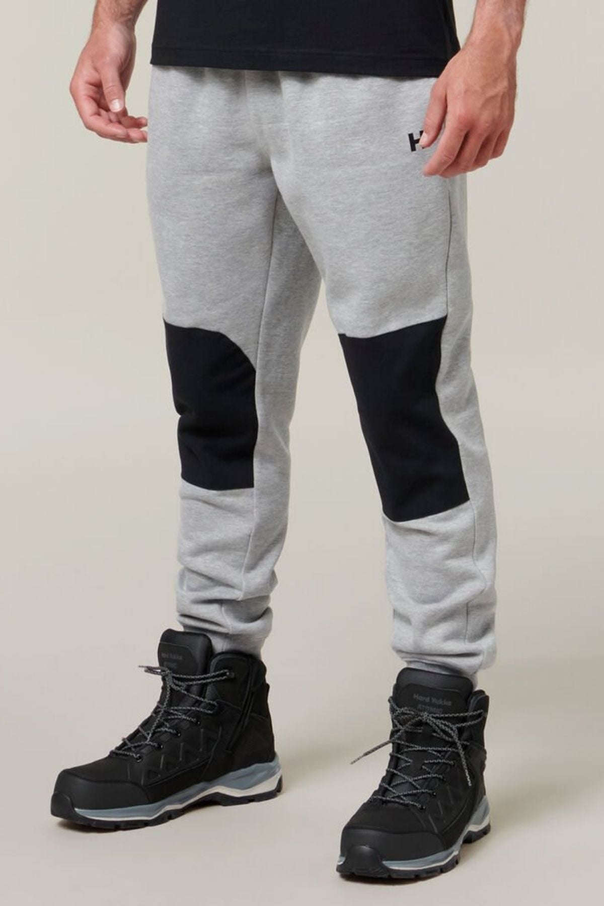 A person is shown wearing Hard Yakka Xtreme Jogger pants with reinforced knee patches and black hiking boots. The grey joggers, featuring a small logo on the upper left thigh, boast a soft fleece lining for added comfort. The individual stands against a plain background.
