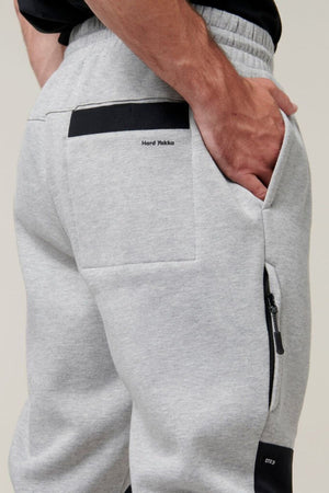 Close-up of a person wearing Hard Yakka Xtreme Jogger track pants in gray, featuring black accents and reinforced knee patches. The person has their right hand in the zippered right pocket. The brand name "Hard Yakka" is visible on the back pocket. The top is not fully visible.