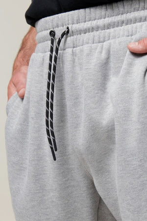 A close-up of a person wearing light gray Hard Yakka Xtreme Jogger pants, showcasing the elastic waistband with a black and white drawstring. The person's right hand is partially visible, tucked into the pants' pocket. These joggers feature reinforced knee patches and soft fleece lining. The background is a neutral, light gray color.