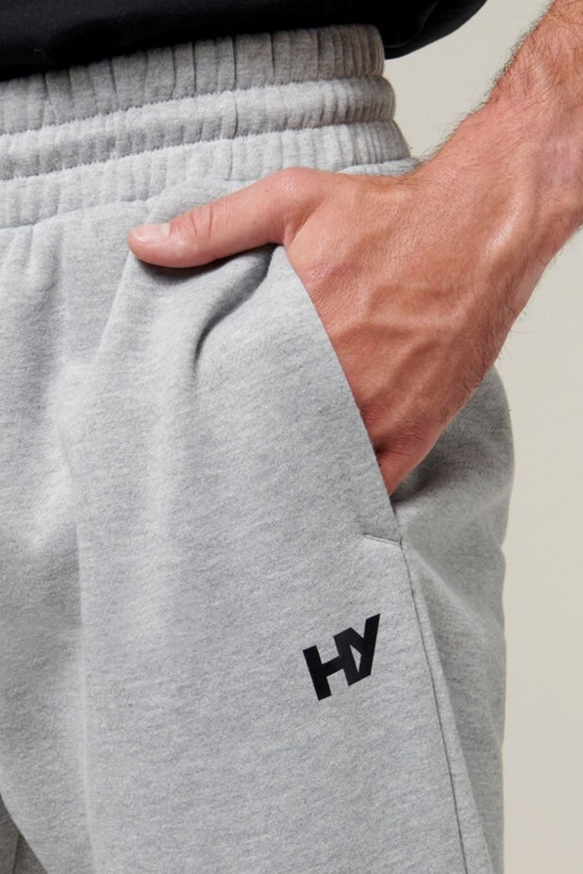 A close-up of a person wearing light gray Hard Yakka Xtreme Joggers with their right hand placed in the pocket. The joggers feature an elastic waistband, a small black "HY" logo on the left thigh, and reinforced knee patches. The background is plain and neutral.