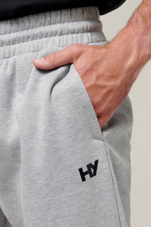 A close-up of a person wearing light gray Hard Yakka Xtreme Joggers with their right hand placed in the pocket. The joggers feature an elastic waistband, a small black "HY" logo on the left thigh, and reinforced knee patches. The background is plain and neutral.