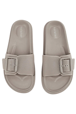 The Holster Laze sandals are light beige vegan slides with wide perforated straps, large buckles, and "holster" printed on the insole, shown from above.