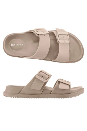 The Holster Soleseeker features beige vegan slip-on sandals with two wide straps and rectangular buckles. One sandal is shown top view, the other side view, both displaying the "holster" branding on the footbed.