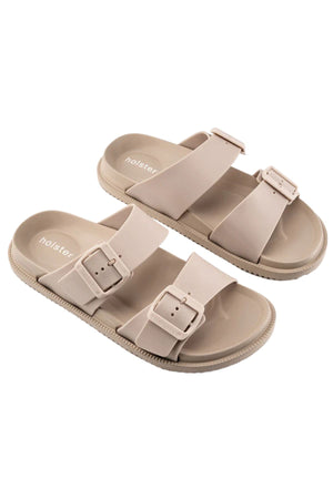 The Holster Soleseeker is a pair of beige vegan slide sandals with two adjustable buckle straps, a textured sole, arch support, and "holster" branding on the insole.