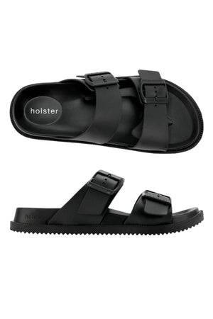 The Holster Soleseeker is a pair of black vegan slip-on sandals with two adjustable buckle straps, flat ridged soles, and "holster" branding on the insole, shown from the top and side views.