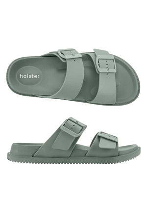 The Holster Soleseeker is a pair of sage green vegan slide sandals with two adjustable buckle straps and a textured waterproof sole. Shown from top and side angles, "holster" is visible on the footbed.