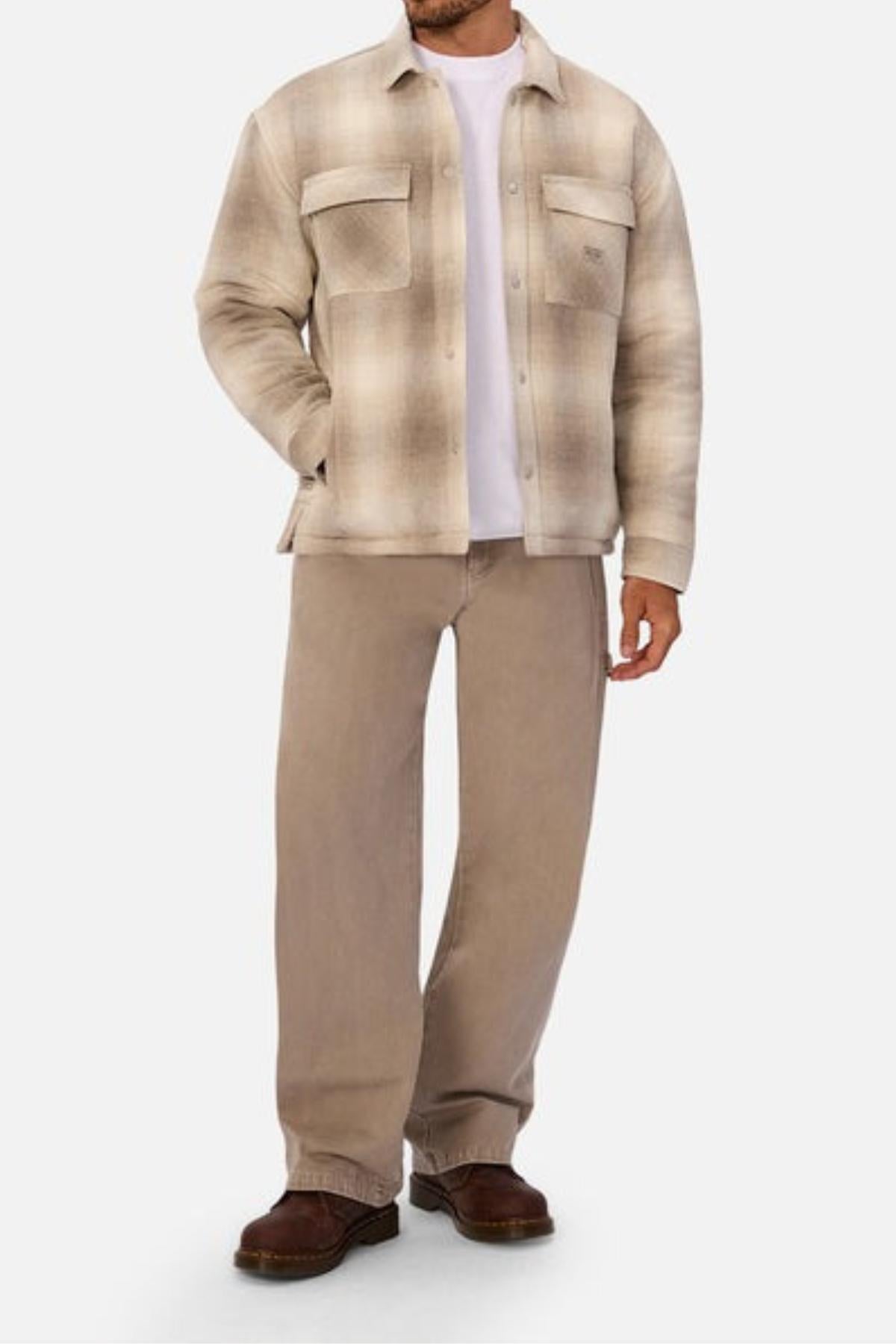 A man stands with one hand in his pocket against a plain white background, wearing the Industrie The Highland Jacket in beige and brown plaid over a white t-shirt, loose tan pants, and brown shoes.
