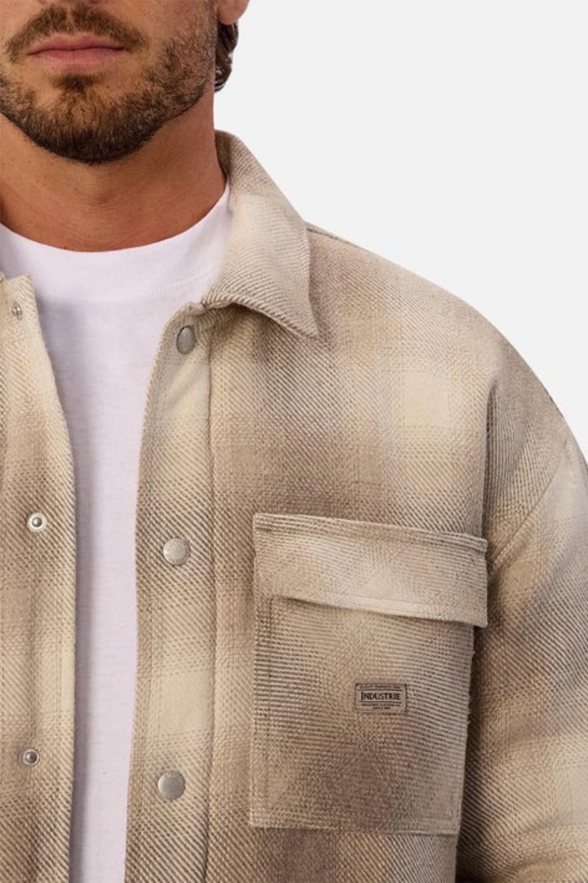 A man wears a light beige plaid button-up shirt with a large chest pocket over a white tee, shown from shoulders to mid-chest against a plain background—ideal for layering under your Industrie The Highland Jacket on outdoor adventures.