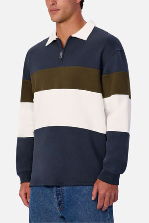 A man wearing the Industrie The Stanstead Tracktop, a contemporary color-blocked pullover with navy, olive green, and white stripes plus a half-zip collar, paired with blue jeans—perfect for casual style.