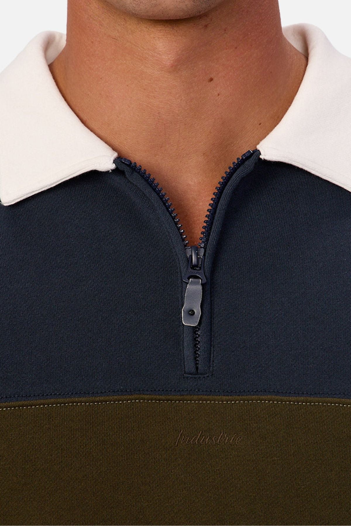 Close-up of a person wearing the Industrie The Stanstead Tracktop in dark blue and olive green with a quarter-zip and white collar, highlighting its contemporary style. Only their lower face and upper chest are visible.