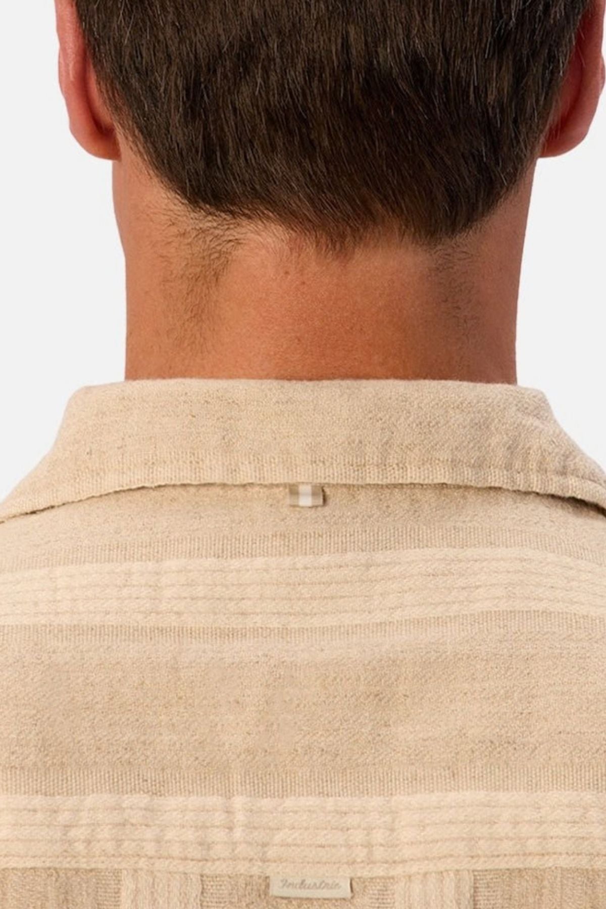 The image shows the back of a person’s neck and head, wearing the Industrie The Ardenza Short Sleeve Shirt in beige. This shirt features a textured fabric, a camp collar, short sleeves, and a small loop detail at the upper back below the collar.