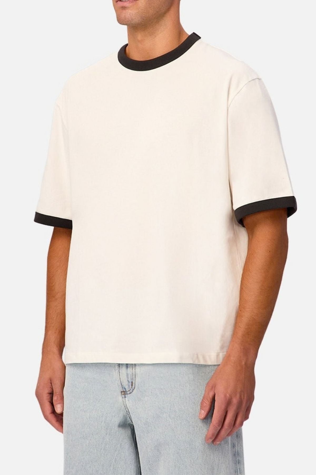 A person wearing the Industrie The Hudson Ringer Tee—a short-sleeve, off-white shirt featuring black trim on the classic crew neckline and sleeves—paired with light blue jeans, stands against a plain light background.