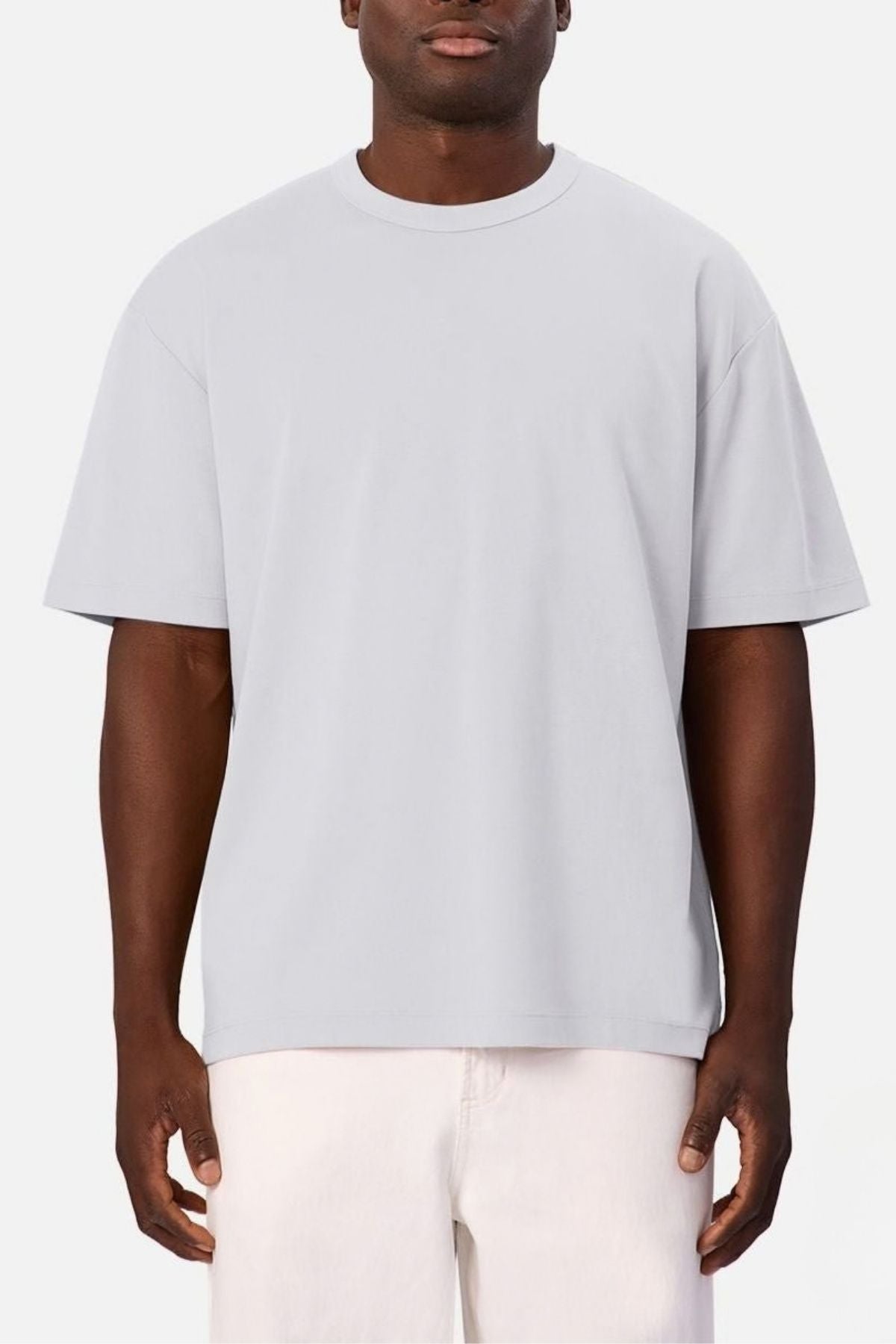 Wearing a relaxed-fit Industrie Luxe Del Sur Tee in light gray—crafted from 100% cotton—a person pairs it with light-colored shorts and stands against a plain white background.