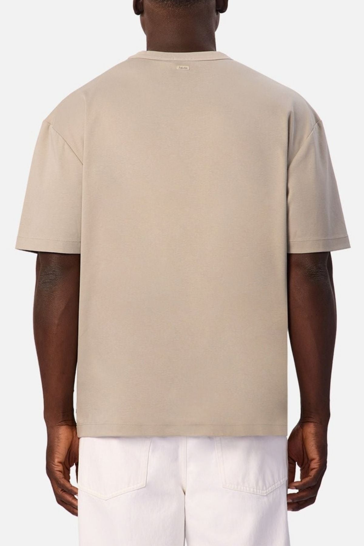 A person is shown from the back wearing the beige Industrie Luxe Del Sur Tee, a 100% cotton relaxed-fit top, paired with white pants and standing against a plain light background.