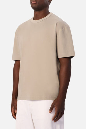 Wearing the Industrie Luxe Del Sur Tee—a loose-fitting beige t-shirt crafted from 100% cotton—paired with white pants, a person stands against a plain white background.