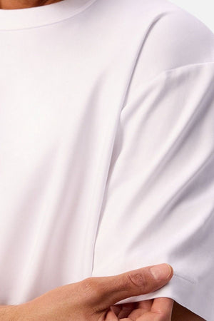 A close-up shows a person wearing the Industrie Luxe Del Sur Tee—a relaxed-fit, 100% cotton t-shirt—holding the sleeve edge between their fingers. Only part of their upper body, arm, and hand are visible.