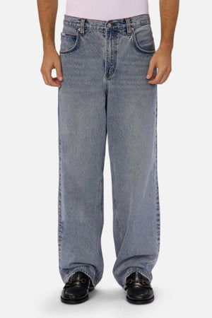 A person wearing Industrie The 80's Jeanโvintage-inspired, loose-fitting wide-leg blue denimโand black loafers stands against a plain white background, with only their lower torso and legs visible.
