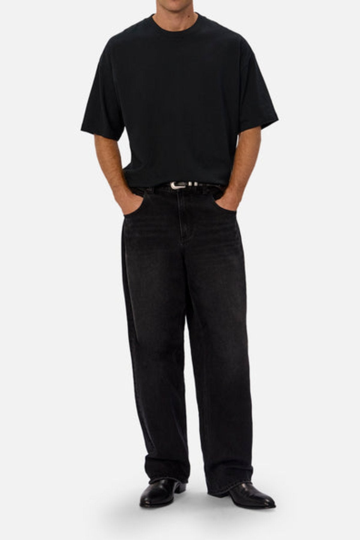 A person in a loose black t-shirt, Industrie The 80's Jean in vintage-inspired durable black denim, a black belt, and black shoes stands hands-in-pockets against a plain light gray background.