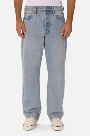 Someone wearing Industrie The 90's Jean in vintage washed light blue, paired with a white shirt and white sneakers, stands facing forward against a plain white background.