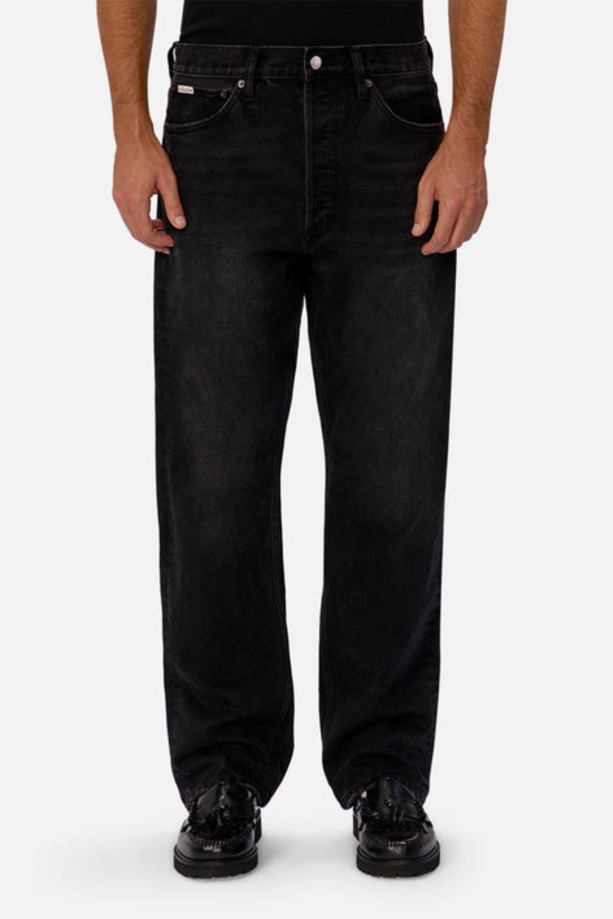 Wearing Industrie The 90's Jean in a relaxed straight leg fit and black color, made from 100% cotton, paired with black shoes and a black top, a person stands against a plain light gray background, shown from shoulders down.