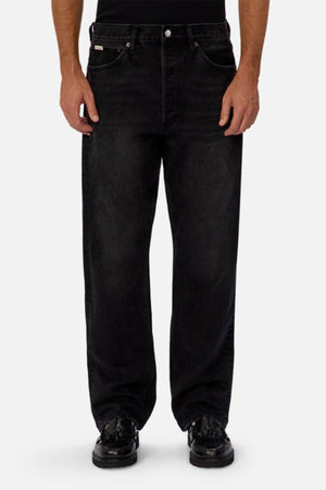 Wearing Industrie The 90's Jean in a relaxed straight leg fit and black color, made from 100% cotton, paired with black shoes and a black top, a person stands against a plain light gray background, shown from shoulders down.