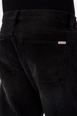 Close-up of the back of a person wearing Industrie The 90's Jean in black vintage washed denim, featuring a visible “Industrie” label above the right pocket. The relaxed straight leg is styled with a black top for a classic look.