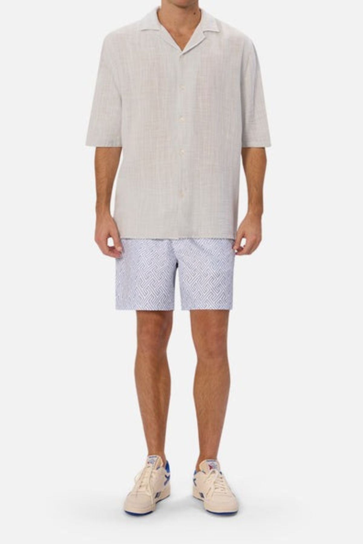 A person wearing a light beige short-sleeve shirt, Industrie The Alvarado Swim Short in white and blue, and cream sneakers with blue accents. The tailored regular fit outfit is casual, summer-inspired, and perfect for quick-drying after a dip.