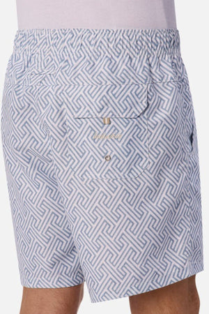 A person wearing the Industrie The Alvarado Swim Short, showcasing a white and light blue patterned fabric with an elastic waistband. These tailored, regular-fit shorts feature a geometric design and include a back pocket with button closure, set against a plain background.