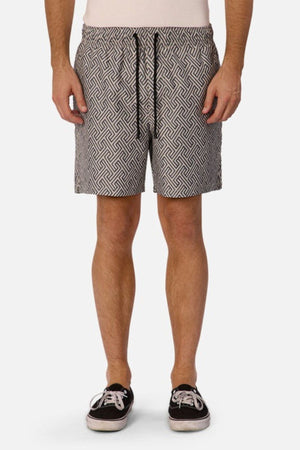 A person wearing Industrie The Alvarado Swim Short featuring a zigzag pattern, a plain light shirt, and black sneakers with white soles stands against a plain white backdrop.