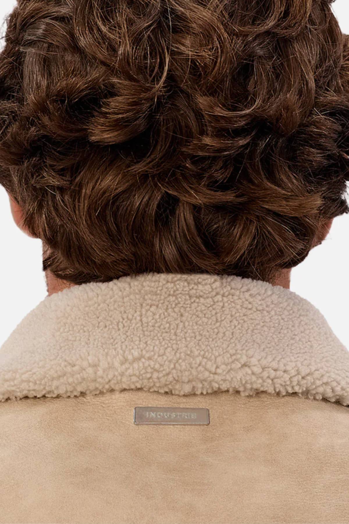A close-up of a person with wavy brown hair wearing the Industrie The Artem Flight Jacket in tan PU leather, featuring a Sherpa-look collar and a small rectangular label at the neckline.