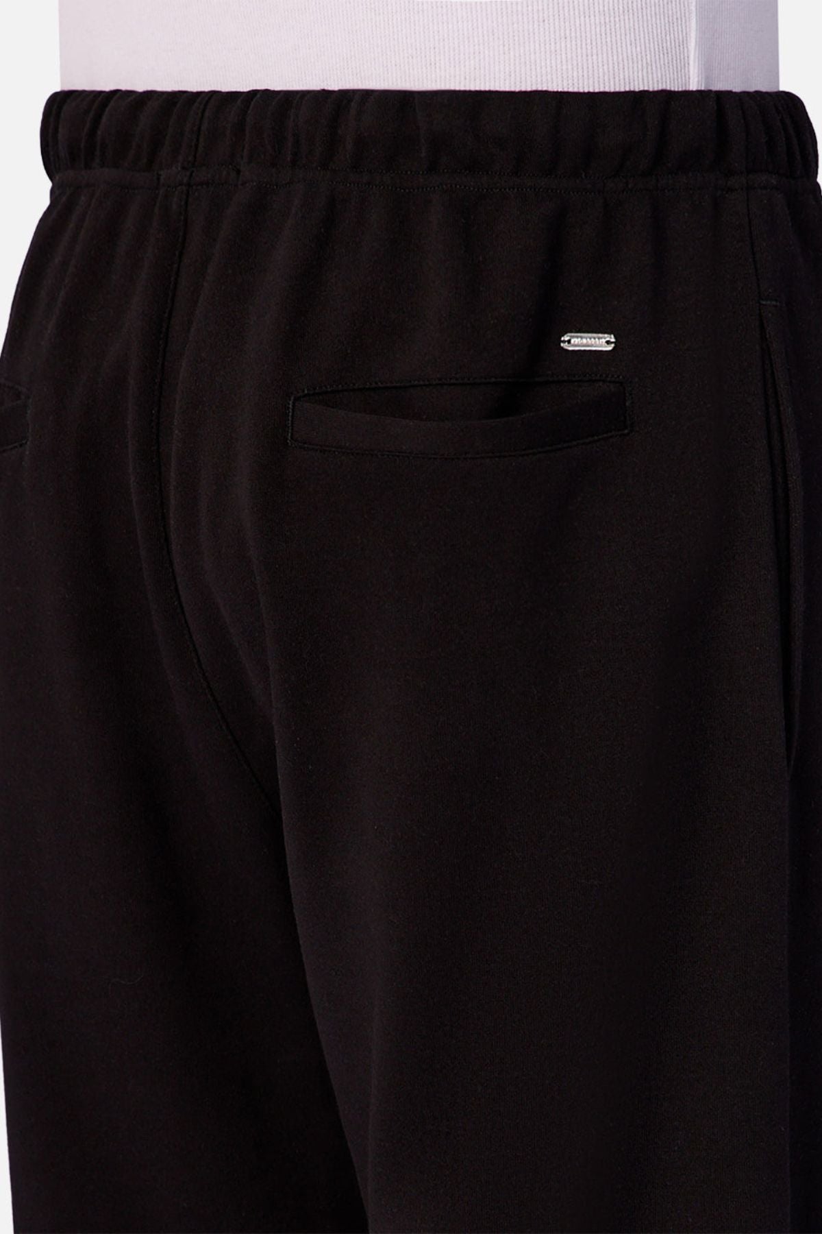 Close-up of a person wearing Industrie The Astoria Lux Tech Trackpant in black with an elastic waistband, back pocket, and metallic logo above the pocket, paired with a white shirt, offering premium comfort and casual style.